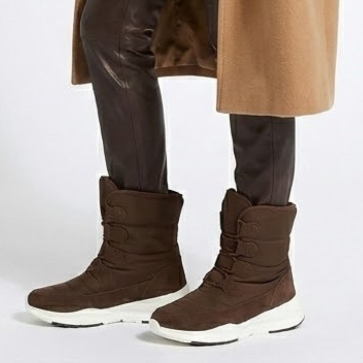 Close-up side profile of the brown Kouvr Gianna snow boot with a white sneaker sole, worn with dark brown leather pants.