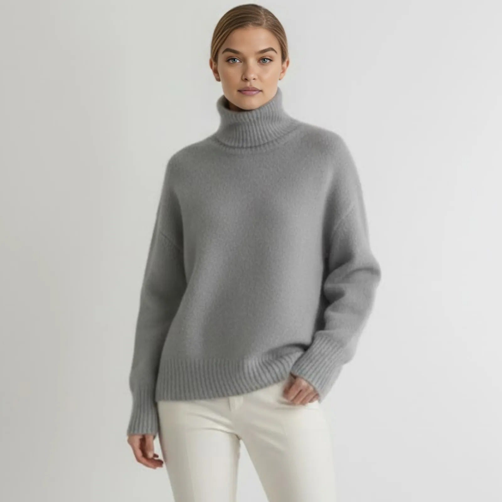 Model facing forward in a heather grey oversized cashmere turtleneck sweater paired with white pants.
