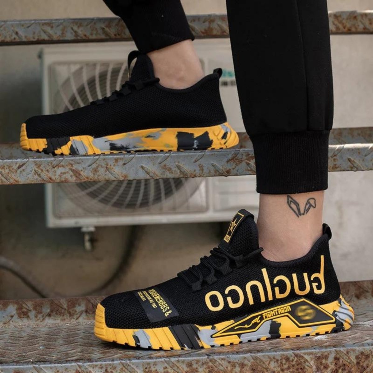 Black and yellow Kouvr sneakers with 'Fugitive' branding worn by a person sitting on a metal surface.