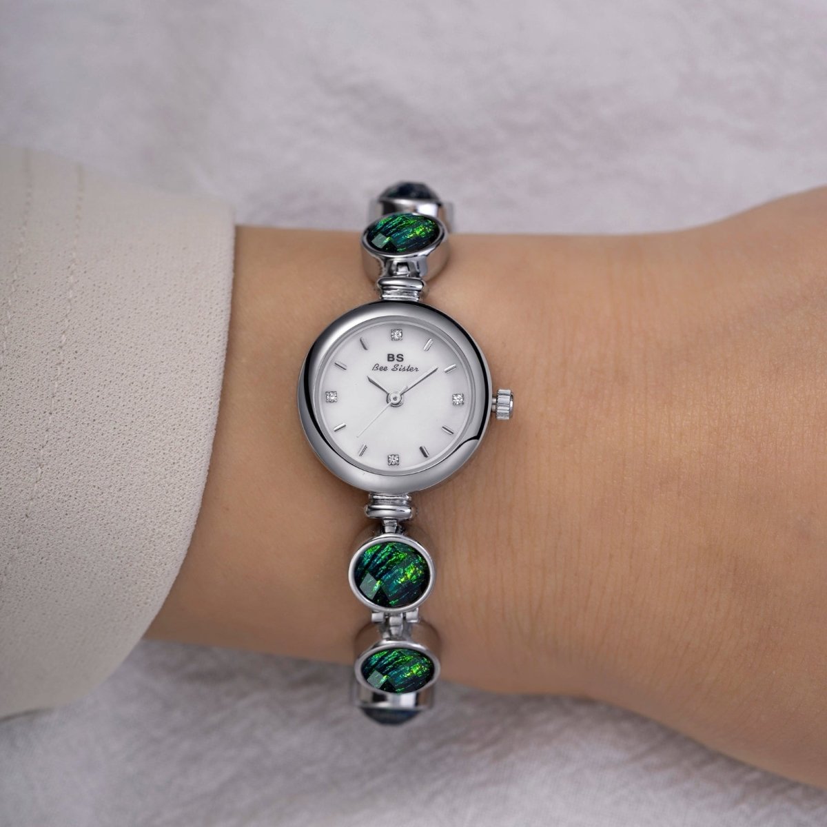 Silver kouvr watch with green gemstones on a wrist against a neutral background