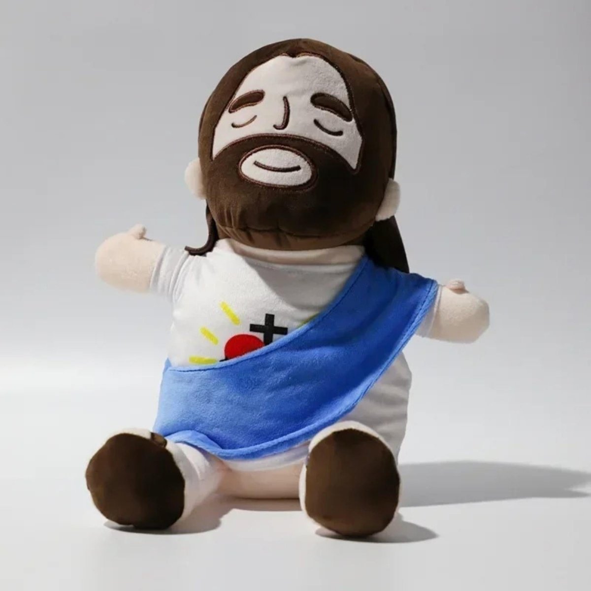Front view of the Kouvr Jesus plush doll with a blue sash, sitting with its arms open in a welcoming gesture against a white background.