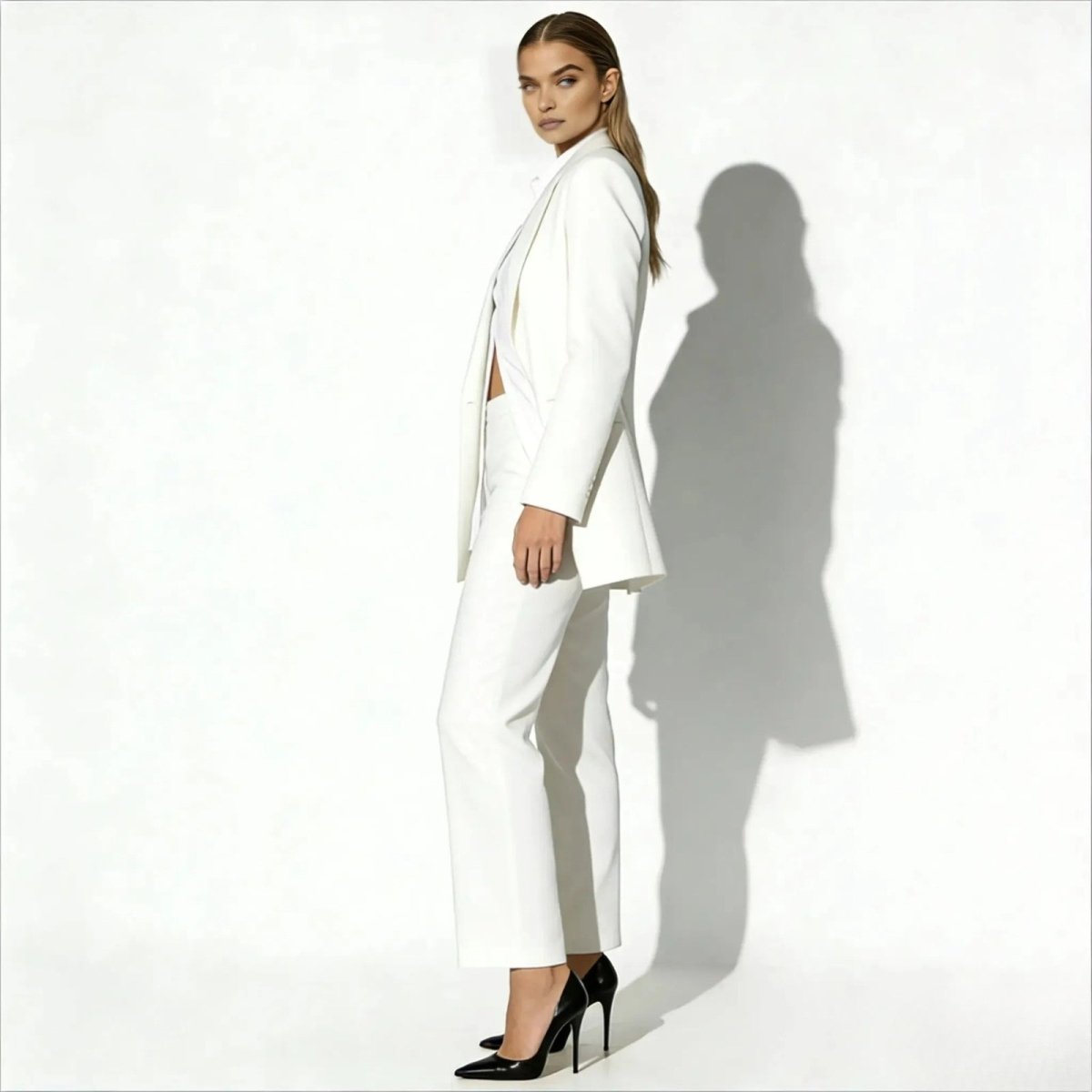 Side view of a model in the Kouvr Kate white suit, showcasing the longline cut of the blazer and the fit of the high-waisted trousers with black heels.
