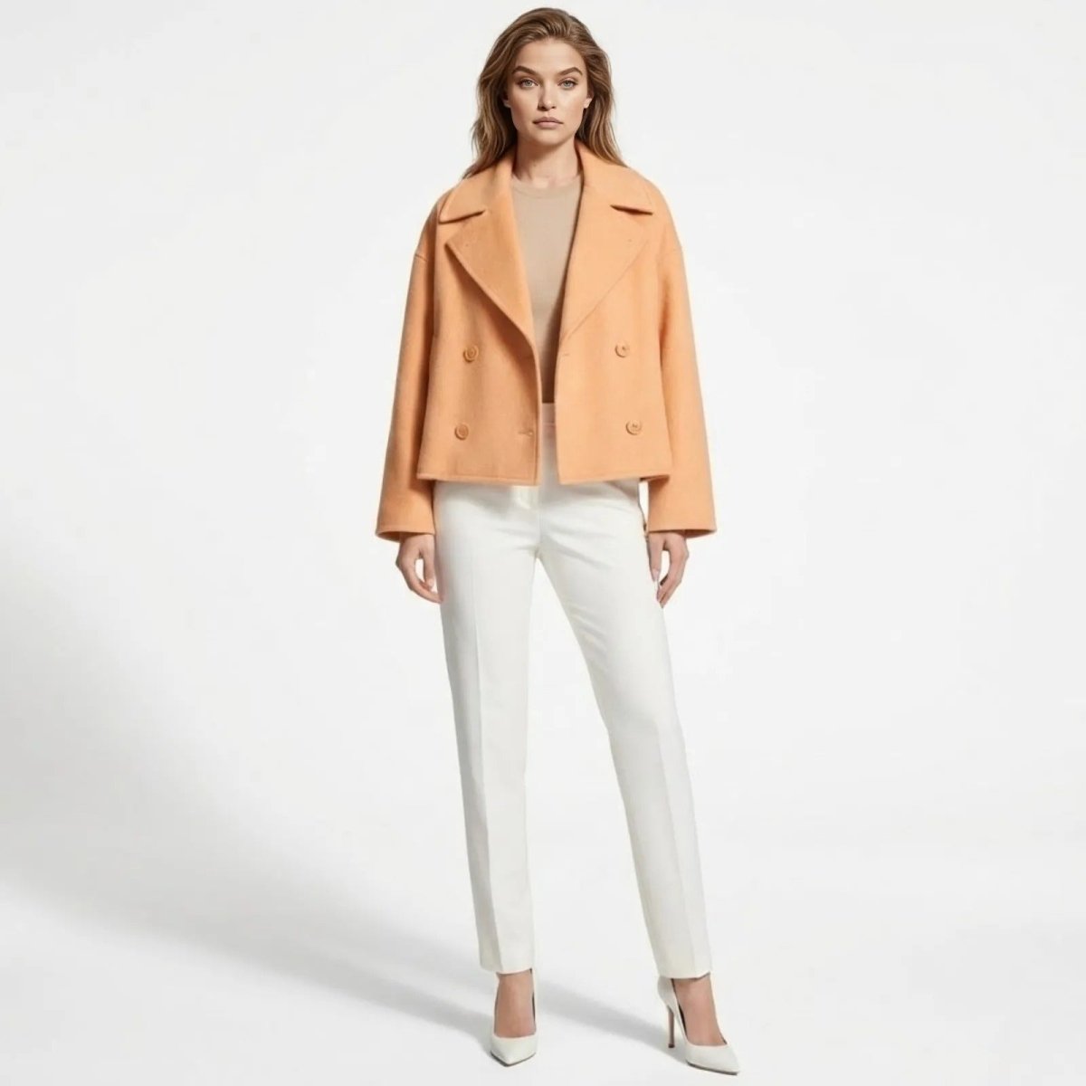 Model wearing the Kouvr Katherine cropped double-breasted wool coat in peach, styled with white trousers, demonstrating its elegant movement and side profile.
