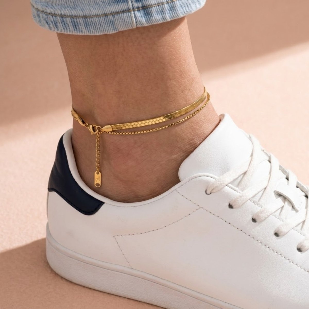 A model wearing the Kouvr layered gold herringbone anklet with classic white sneakers and light-wash denim.