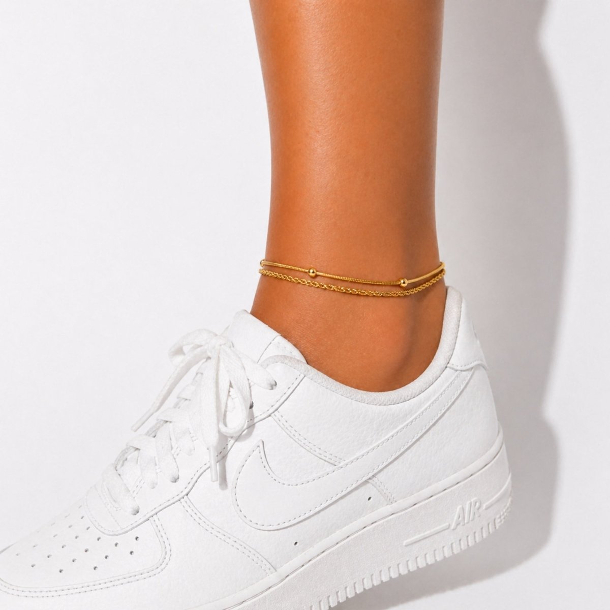 Model wearing the Kouvr double chain gold anklet with classic white sneakers against a white wall.