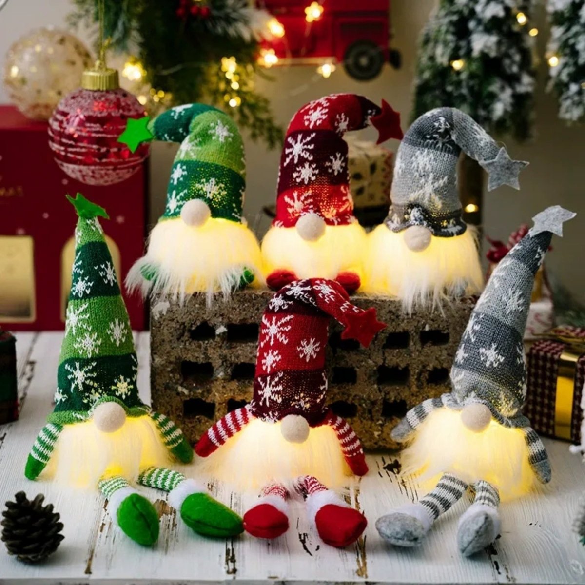 A set of three handcrafted Kouvr Christmas gnomes in red, green, and grey, illuminated with a warm LED glow, arranged on a rustic wooden surface with festive holiday decorations in the background. 