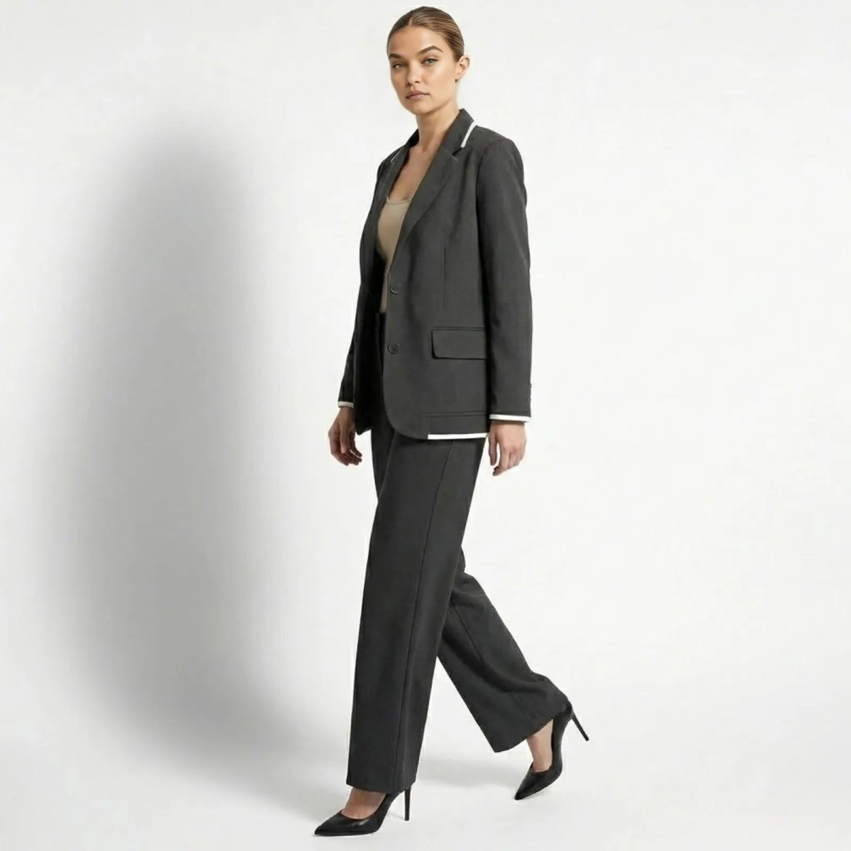 Side profile of a model walking in the Kouvr Lucia Set dark grey wide-leg pant suit, showcasing the fluid drape of the trousers and the tailored fit of the blazer with white cuff details.
