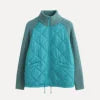 Model wearing the Kouvr Mika Hybrid Bomber Jacket in Aegean Teal, featuring cable-knit sleeves and a diamond-quilted body, paired with light-wash skinny jeans and white sneakers