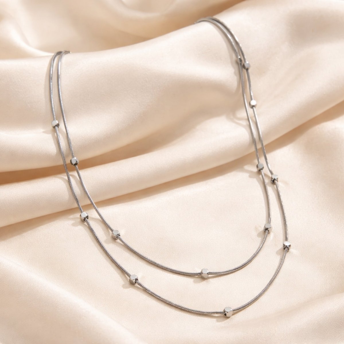 Close up of the delicate Kouvr layered silver station necklace resting on cream-colored silk fabric.