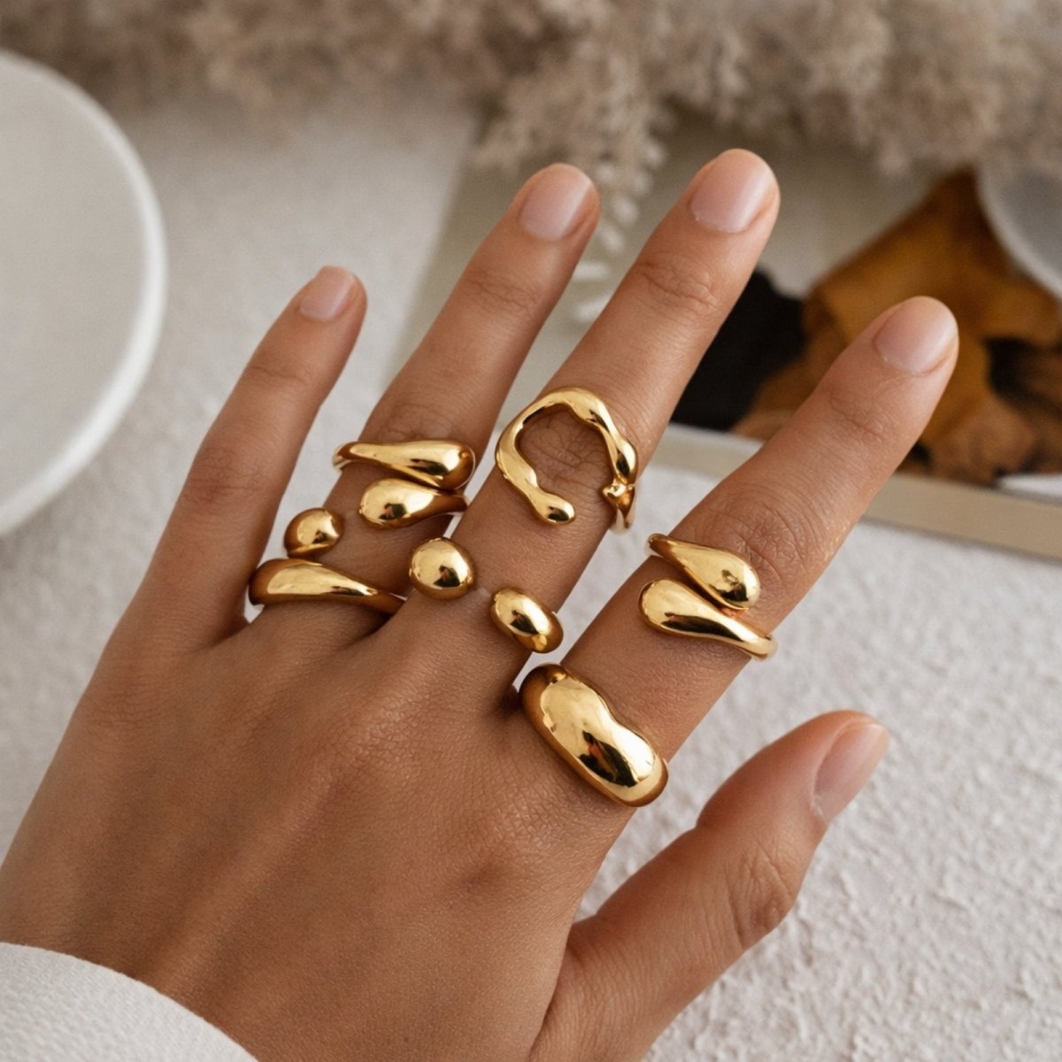 A styled hand displaying multiple chunky gold rings from the Kouvr collection, showing their molten, organic shapes against a soft, neutral background.
