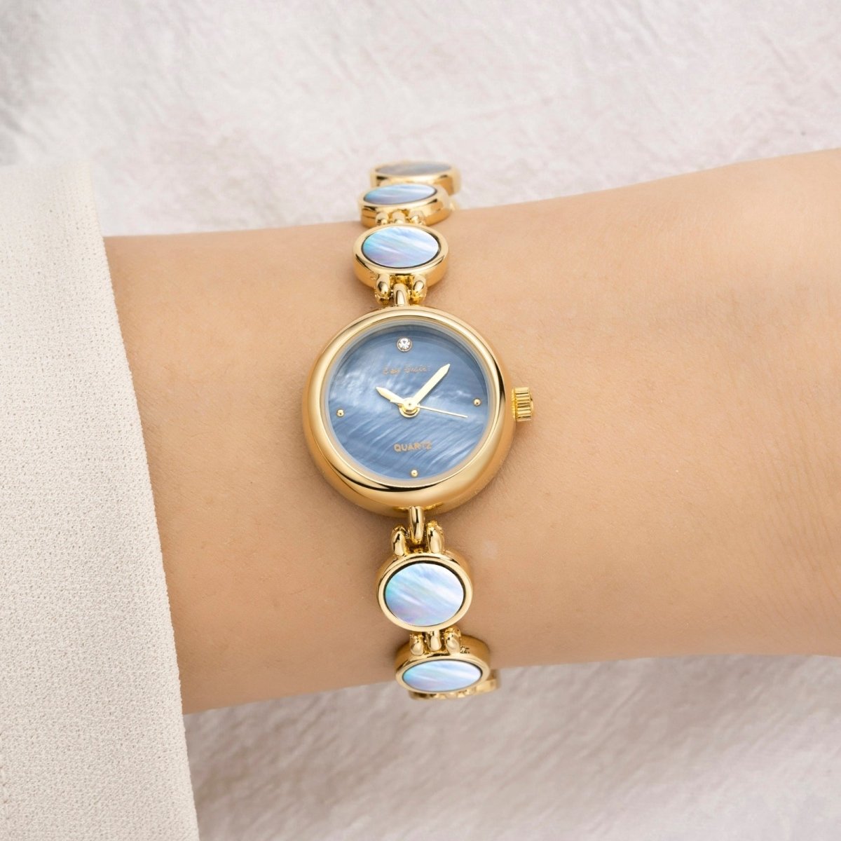 A woman is wearing a Kouvr Moon Gold Faelan gold-tone watch with a white mother of pearl bracelet and dial.