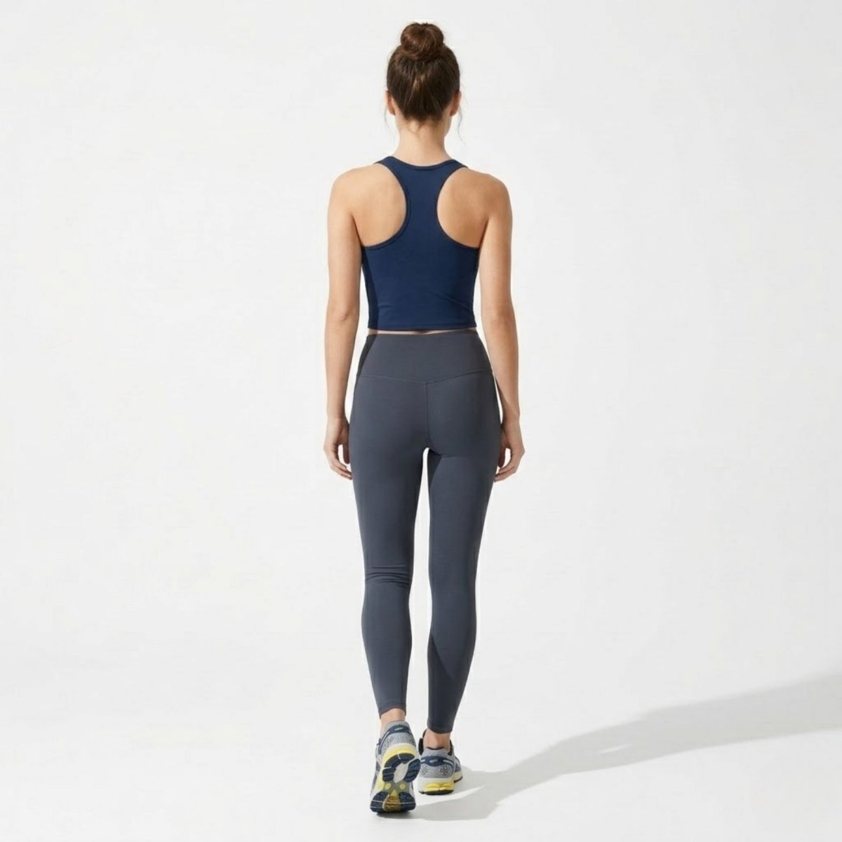 Full body back view of model walking in Kouvr navy blue racerback sports top and grey leggings.