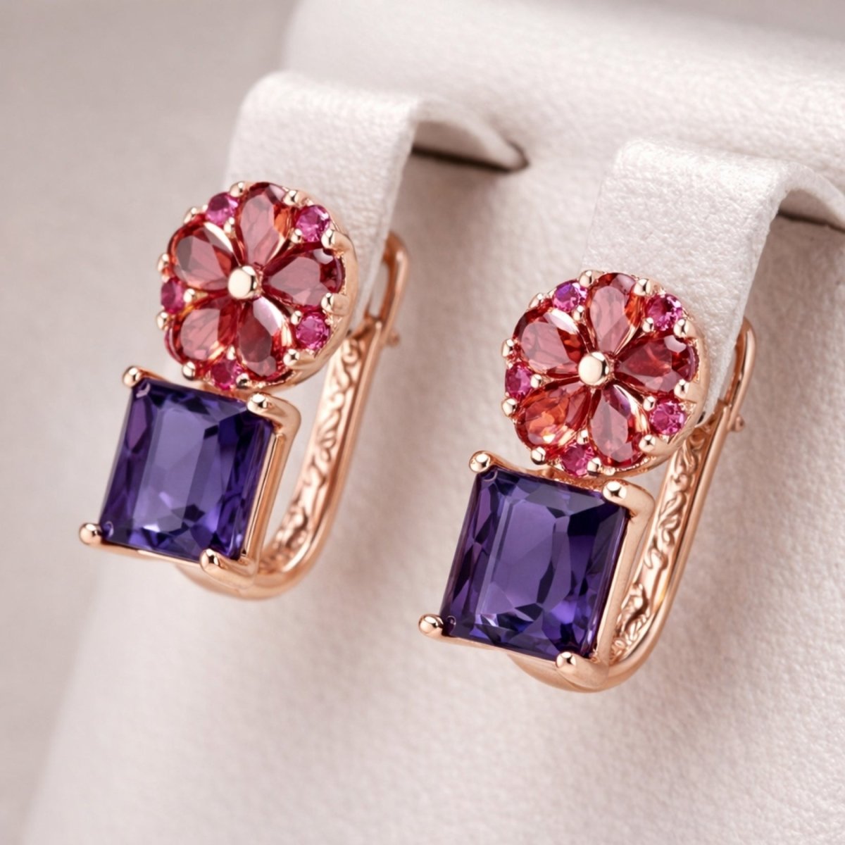 Pair of Kouvr Octivia flower drop earrings with square amethyst and red garnet in 18k rose gold on a white display stand.