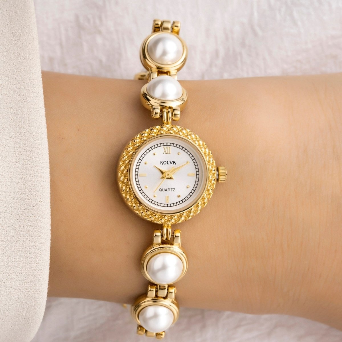 Gold kouvr watch with pearl accents on a wrist against a neutral background