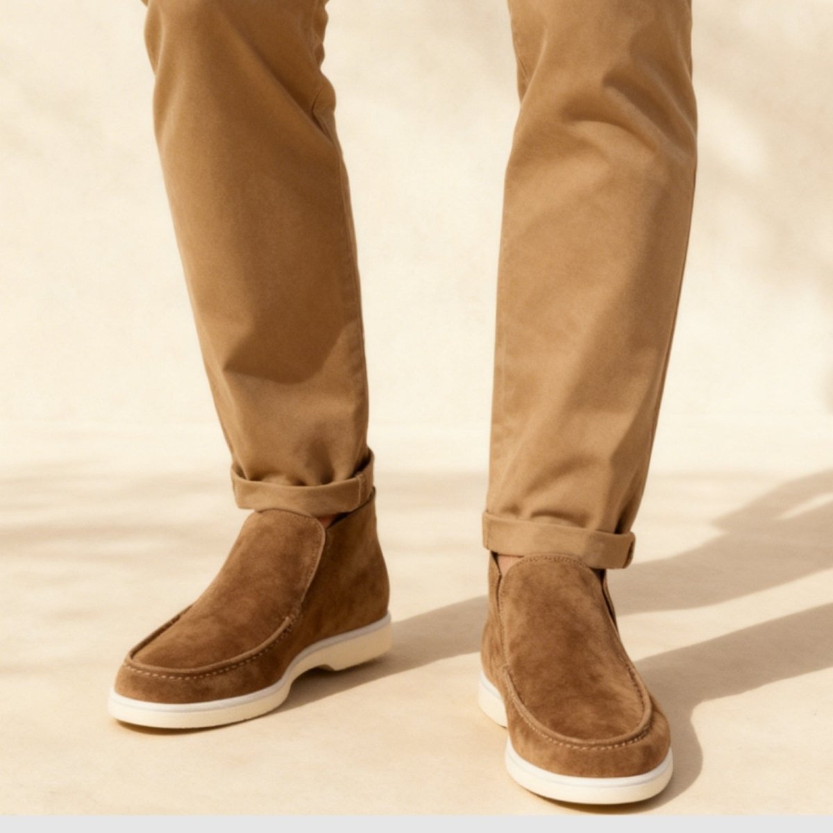 A man's feet wearing brown tan suede slip-on loafer boots with cuffed khaki chinos, viewed from the front on a light-colored floor. 