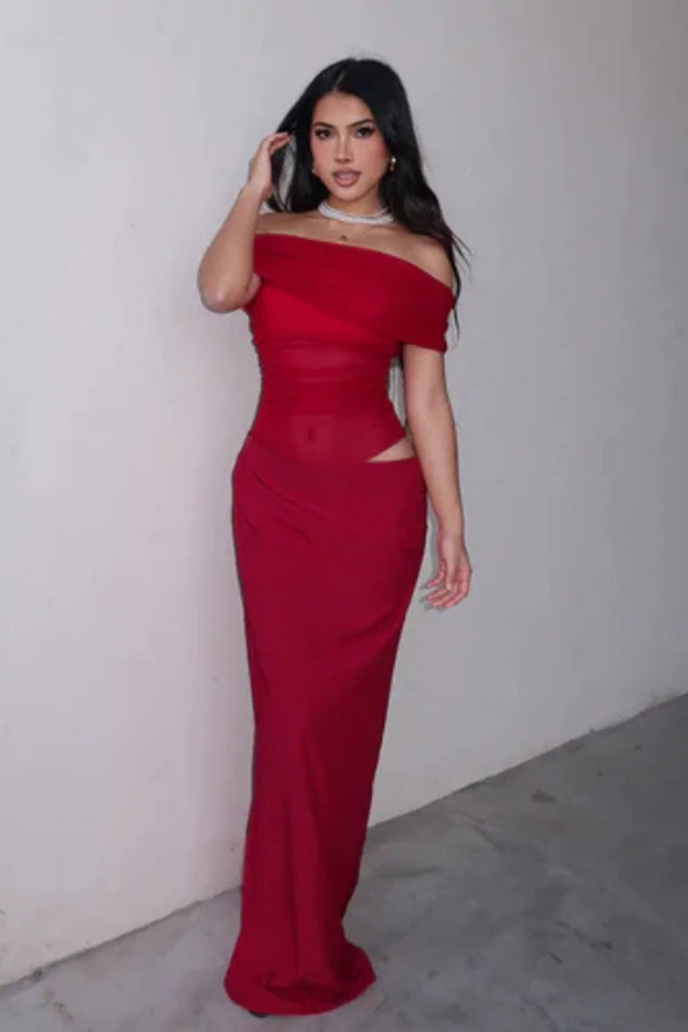 Front view of a woman wearing the Kouvr red off-shoulder formal gown with a sheer waist and hip cutout