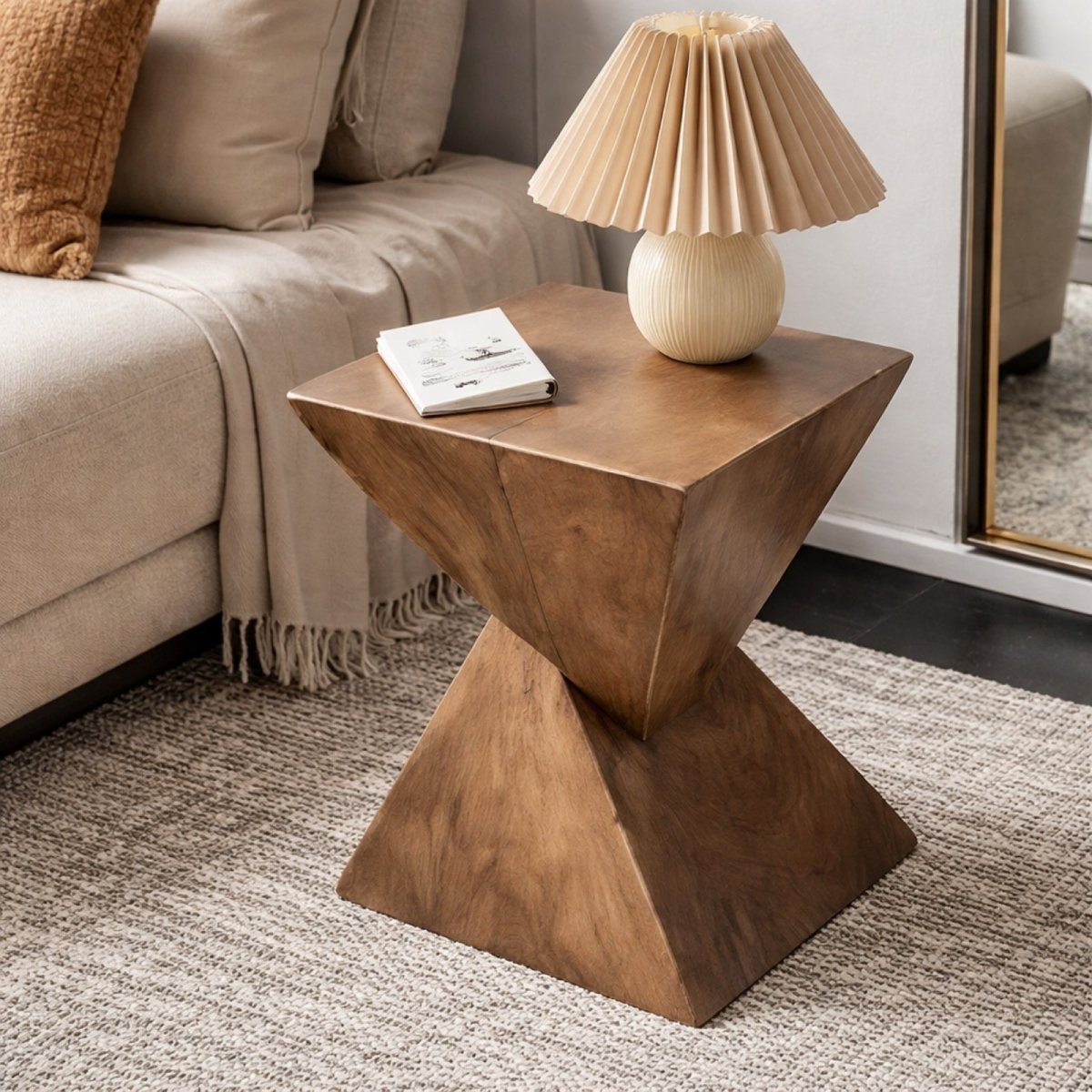 A dark wood finish geometric accent table serving as a nightstand next to a sofa, holding a book and a pleated cream-colored table lamp.