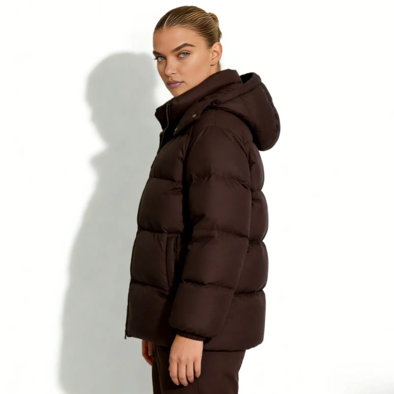 Side profile of a woman in a dark brown quilted down puffer jacket by Kouvr, showing its hip-length and hood volume.