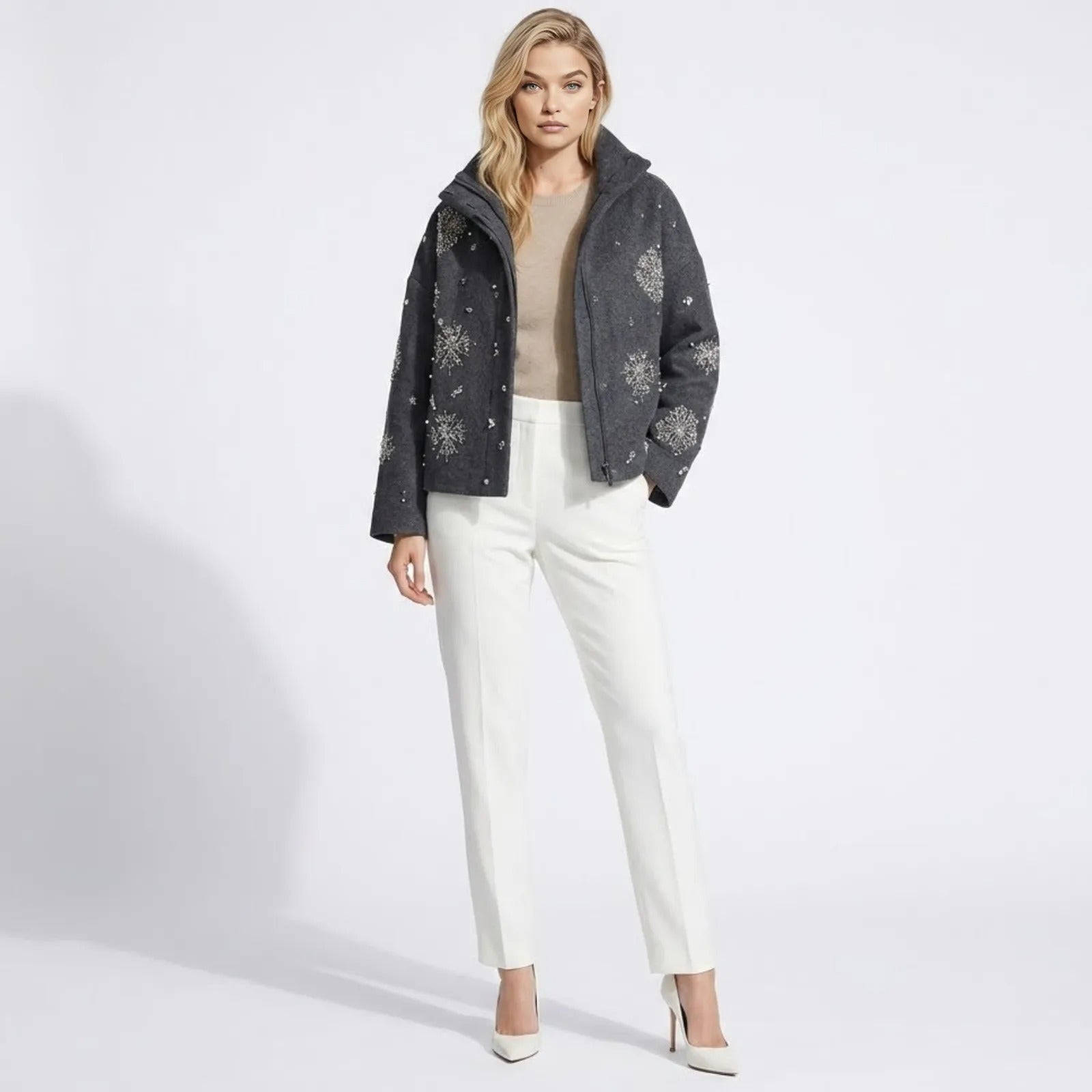 Female model wearing the Kouvr dark grey wool jacket with silver crystal snowflake embellishments, paired with white trousers and white heels against a white background.