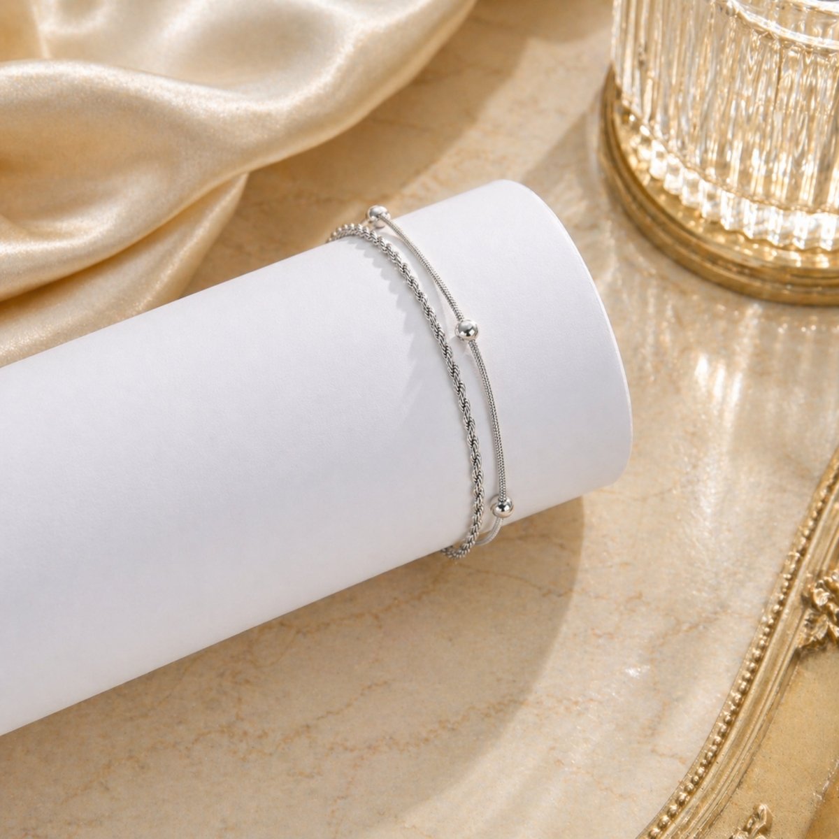 The Kouvr silver layered anklet displayed on a white cylinder with elegant gold and cream decor.