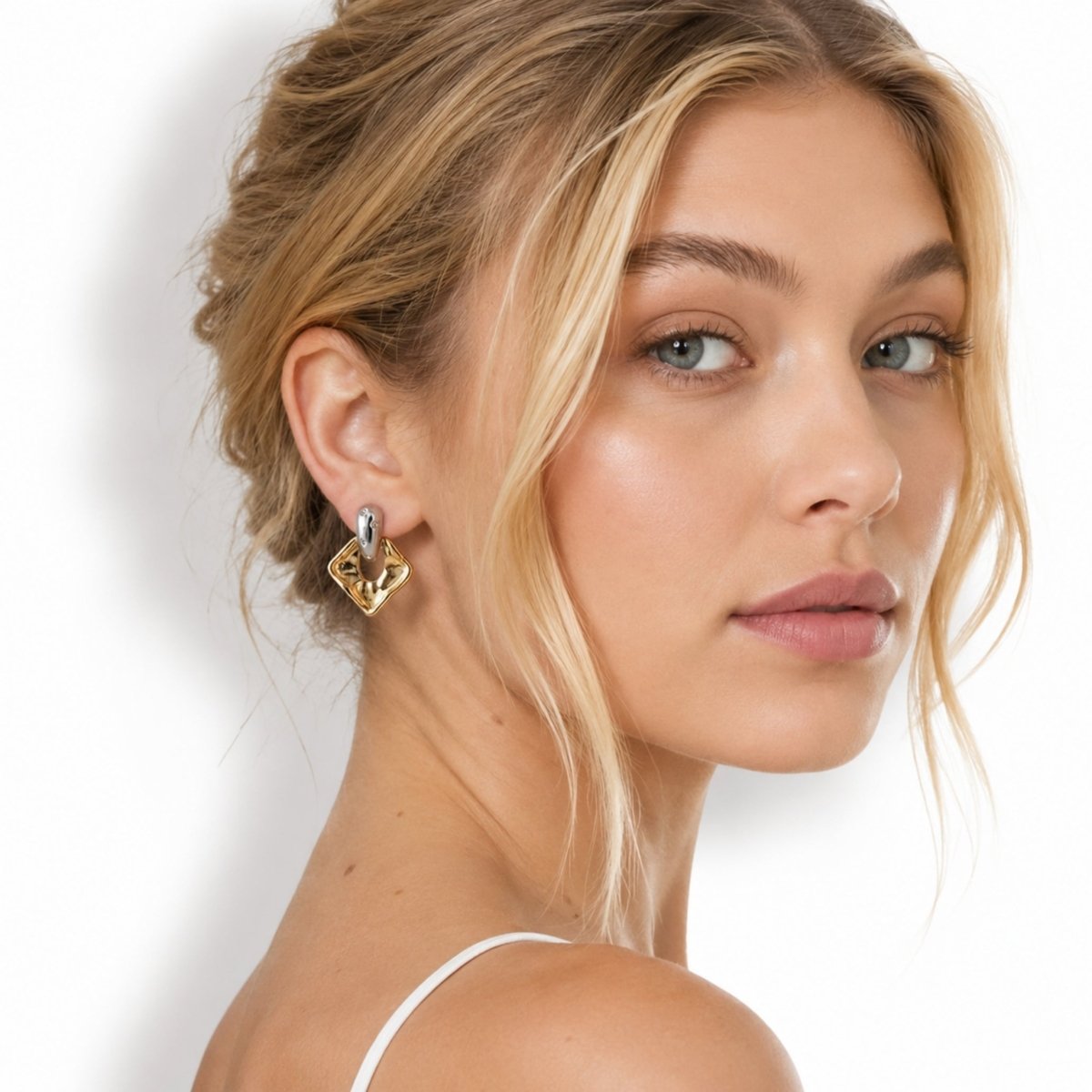 Blonde female model looking over her shoulder wearing Kouvr sculptural two-tone gold and silver drop earrings with a white strap top.
