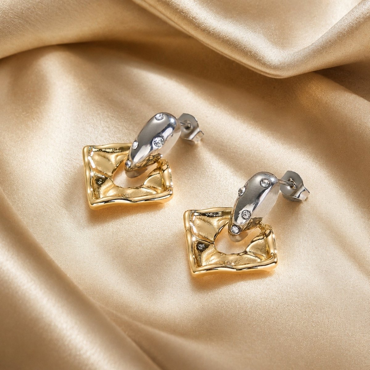 Pair of Kouvr two-tone metallic statement earrings with embedded crystals resting on luxurious draped gold silk fabric.