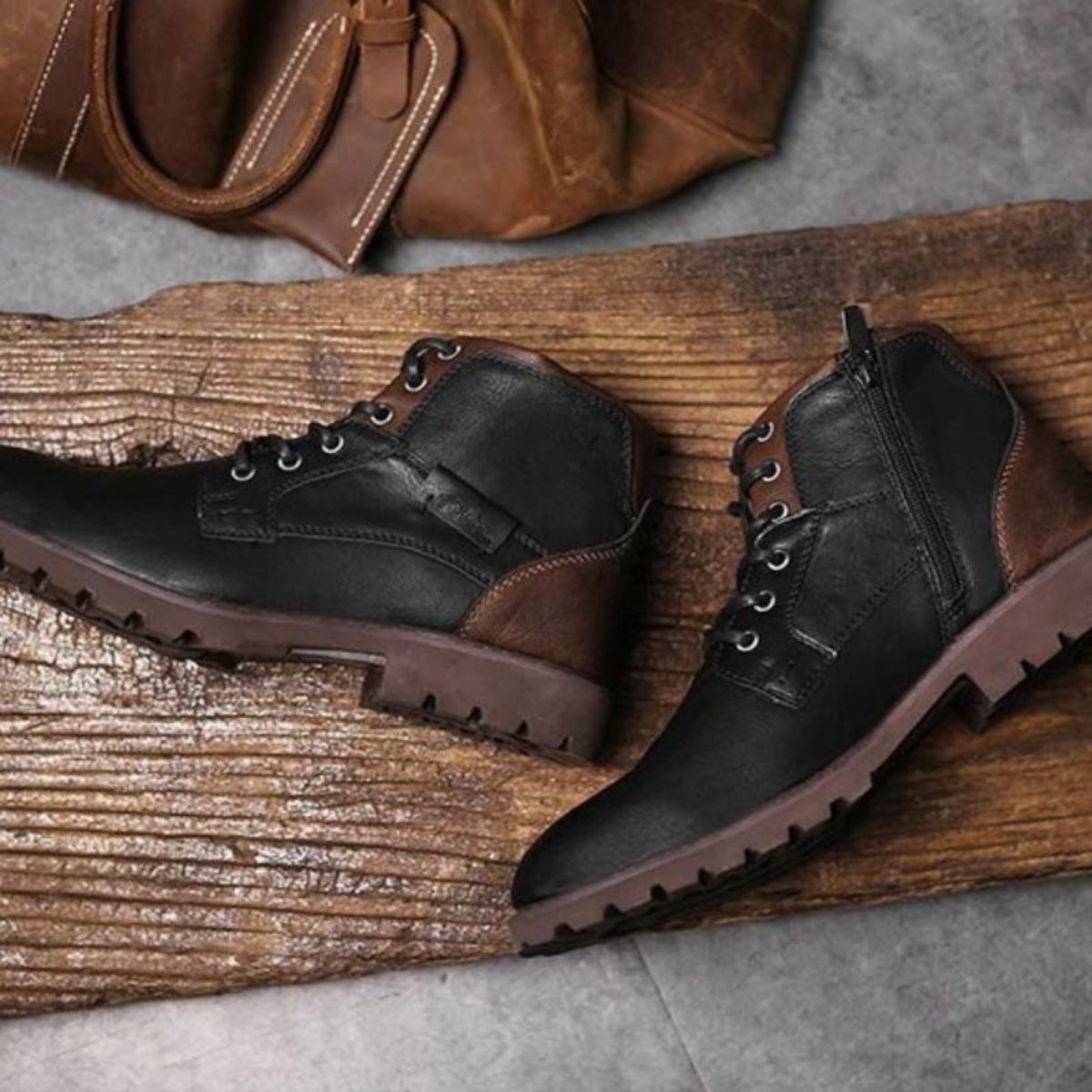 Pair of black leather boots on a wooden surface with a brown leather bag in the background.