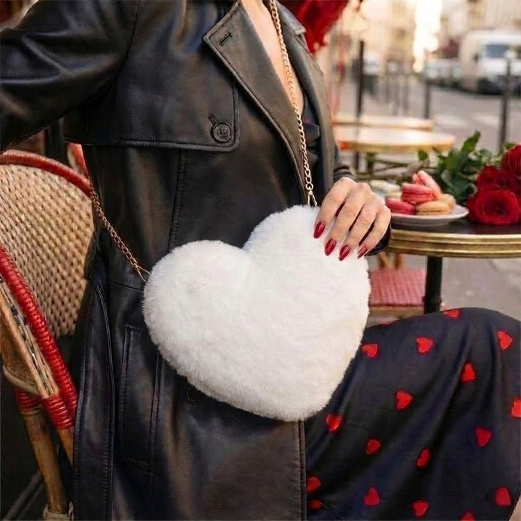 Woman in a black leather trench coat and heart-print dress sitting at a Parisian cafe, wearing the Kouvr white fluffy heart-shaped crossbody bag.
