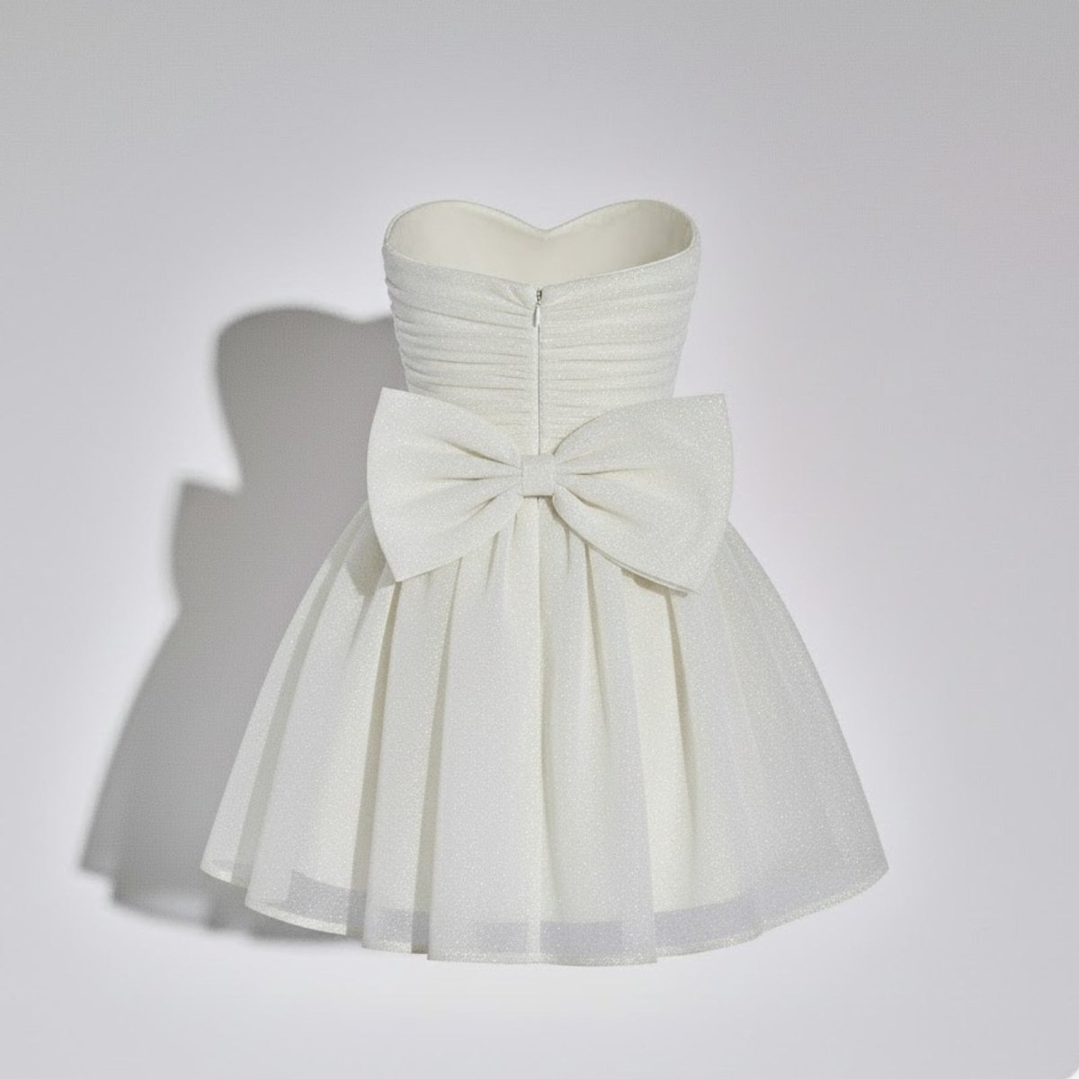 Close-up product shot of the back of a white strapless mini dress, highlighting the oversized architectural bow detail and central zipper.
