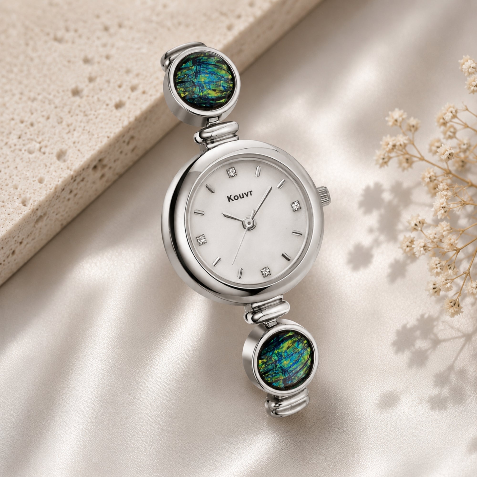 Kouvr silver women's jewelry watch resting elegantly on shimmering cream silk fabric next to a textured stone block and delicate white dried flowers.