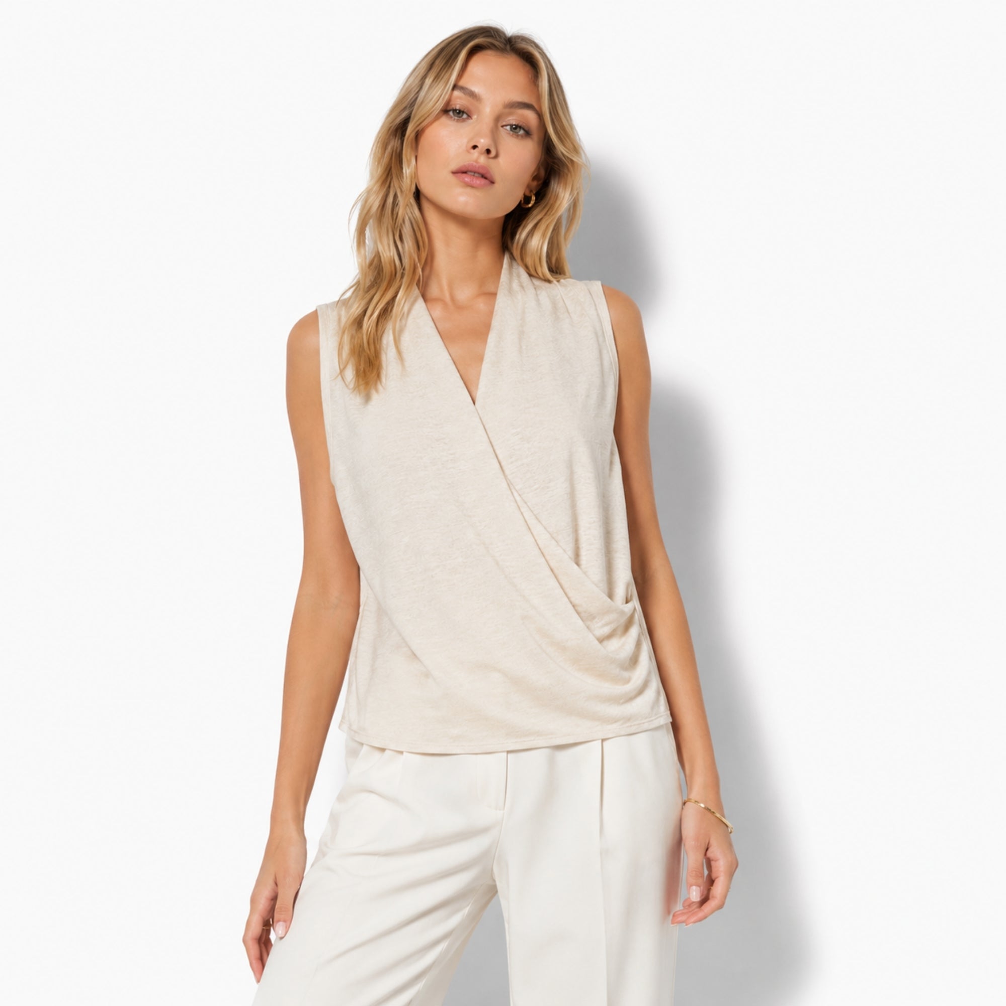 Woman wearing a sophisticated beige sleeveless draped wrap blouse with a v-neckline by Kouvr, styled with tailored cream trousers.