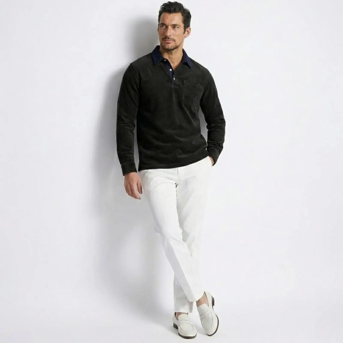 Male model wearing the Kouvr Zain Black Corduroy Long Sleeve Polo Shirt with white trousers, leaning against a white wall.