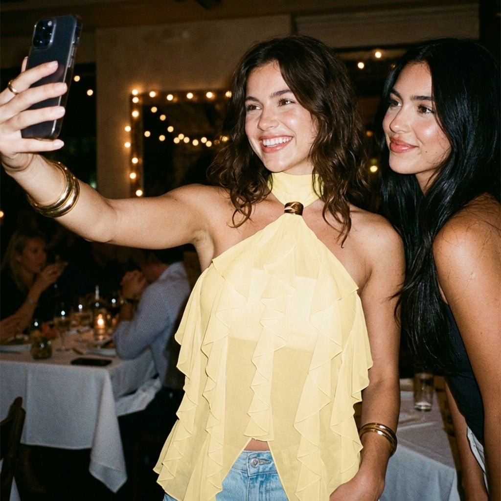 Two women taking a selfie at a dinner party, one wearing the pale yellow sheer ruffle halter top styled with gold bangles.