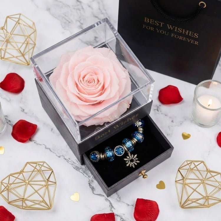 Close-up of a pastel pink eternal rose in a black box with a "Best Wishes" logo, featuring a snowflake charm bracelet in the open drawer.