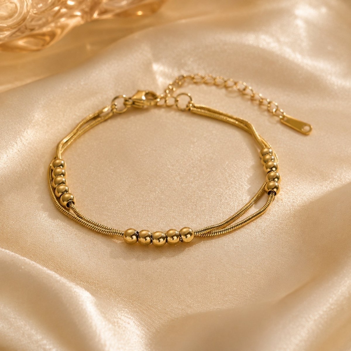 Close up of luxury 18K gold double layered beaded anklet resting on elegant champagne silk fabric.