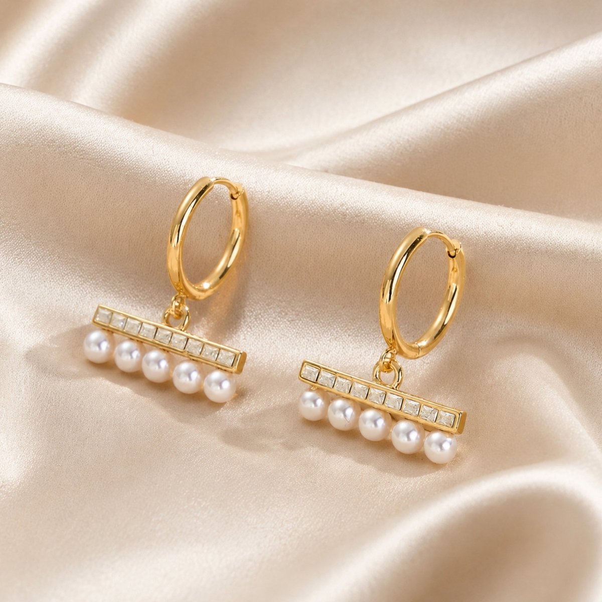 Luxurious gold huggie hoop earrings with luminous pearls and sparkling square crystals resting elegantly on draped champagne silk fabric.