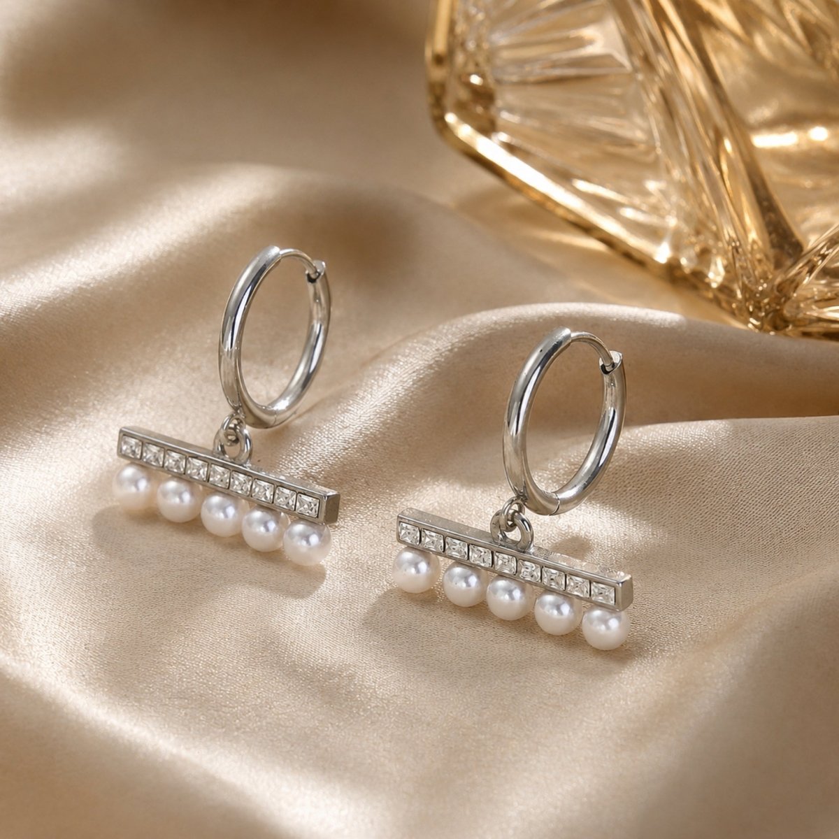 Sophisticated silver pearl and crystal dangle hoop earrings displayed next to vintage textured glass on warm, luxurious beige silk.