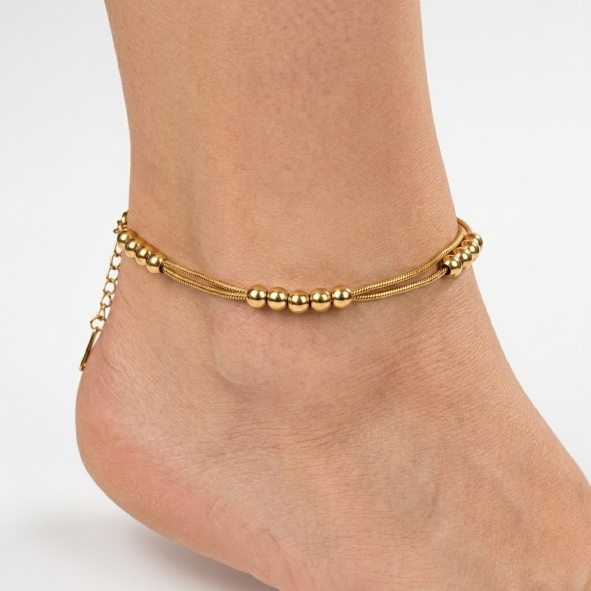 Woman's foot wearing an 18K gold double layered snake chain anklet with delicate metal beads.