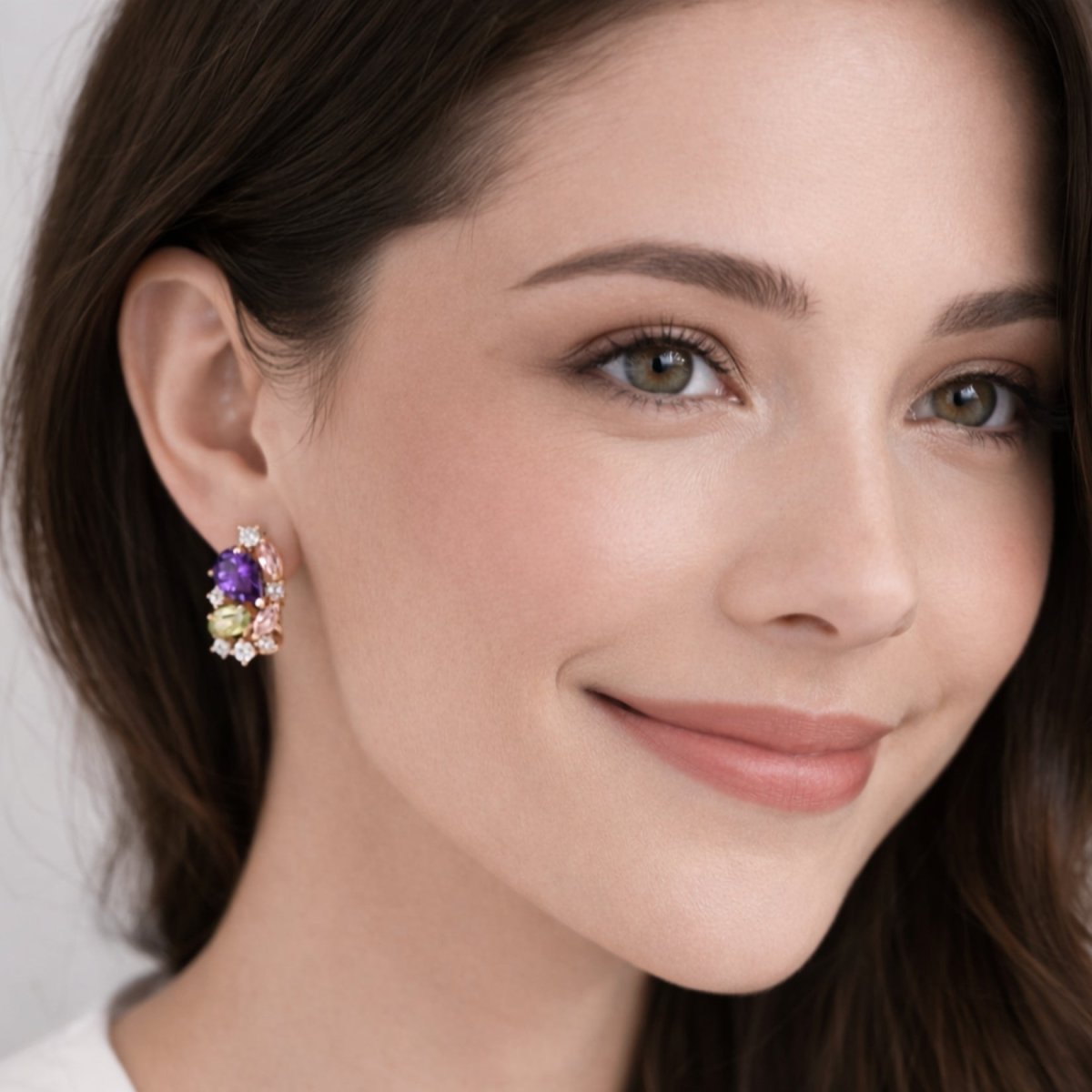 A smiling woman with brown hair wearing the Kouvr Aurora multi-gemstone cluster earrings, showcasing their size and vibrant colors on the ear.