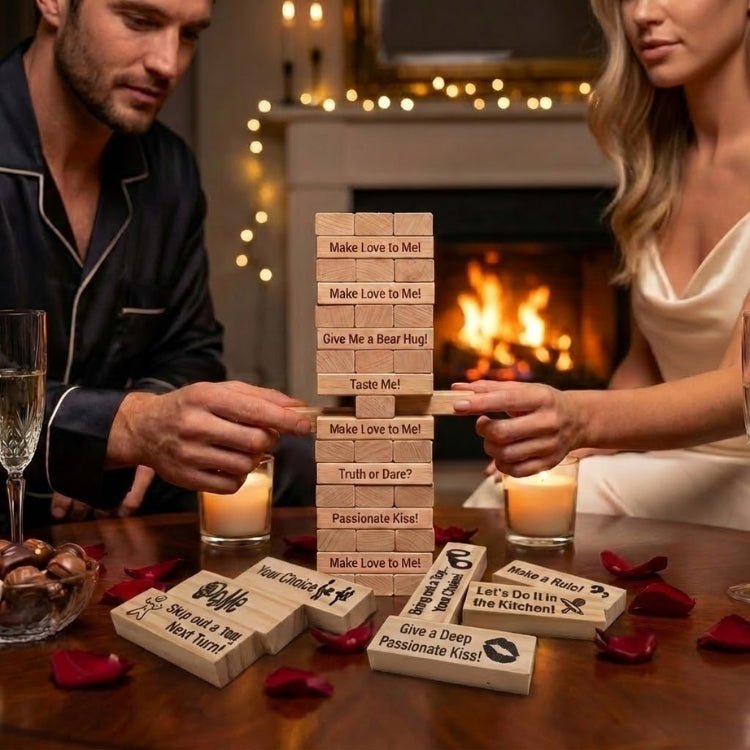 A man and woman in silk loungewear playing a 48-piece naughty wooden block game for adults on a table with rose petals and wine by a cozy fireplace.