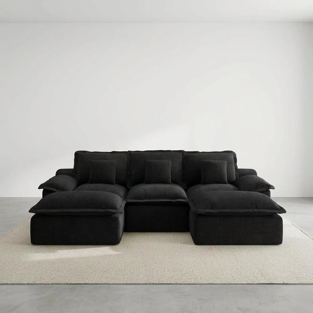Oversized black sectional couch with deep seats and extra pillows placed in a sunny living room near a marble fireplace.