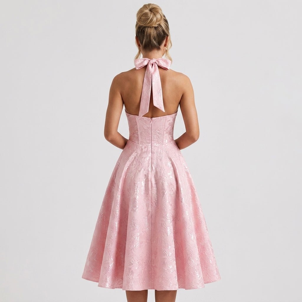 Back view of pink open back formal event dress highlighting large tied halter neck bow and pleated tea-length skirt.
