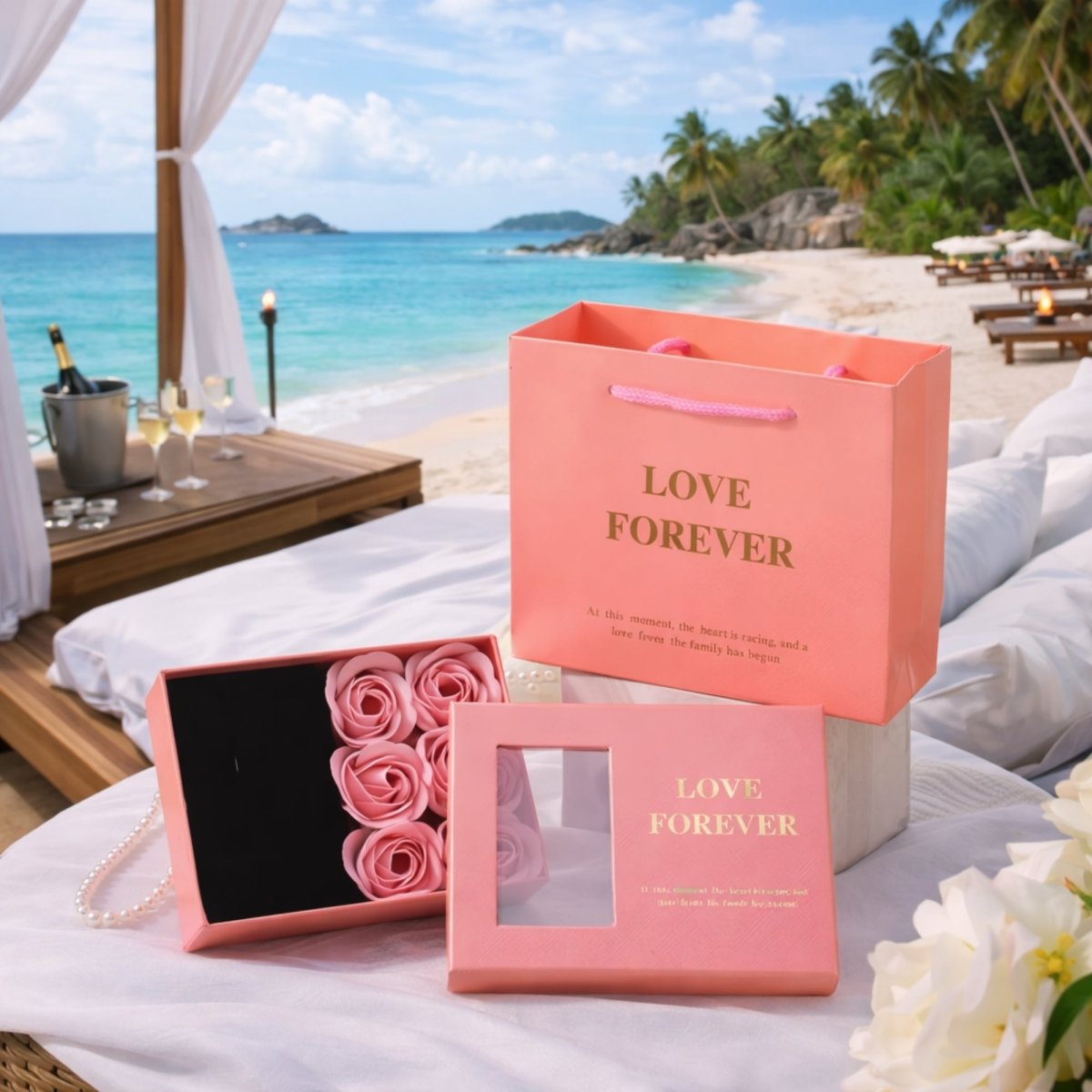 A romantic and beachy pink gift set on a white tablecloth in the Seychelles, featuring a pink 'Love Forever' paper bag and two boxes with six pink preserved roses, a gold necklace with a sunflower pendant, and a pearl bracelet, with ocean and daybed views.