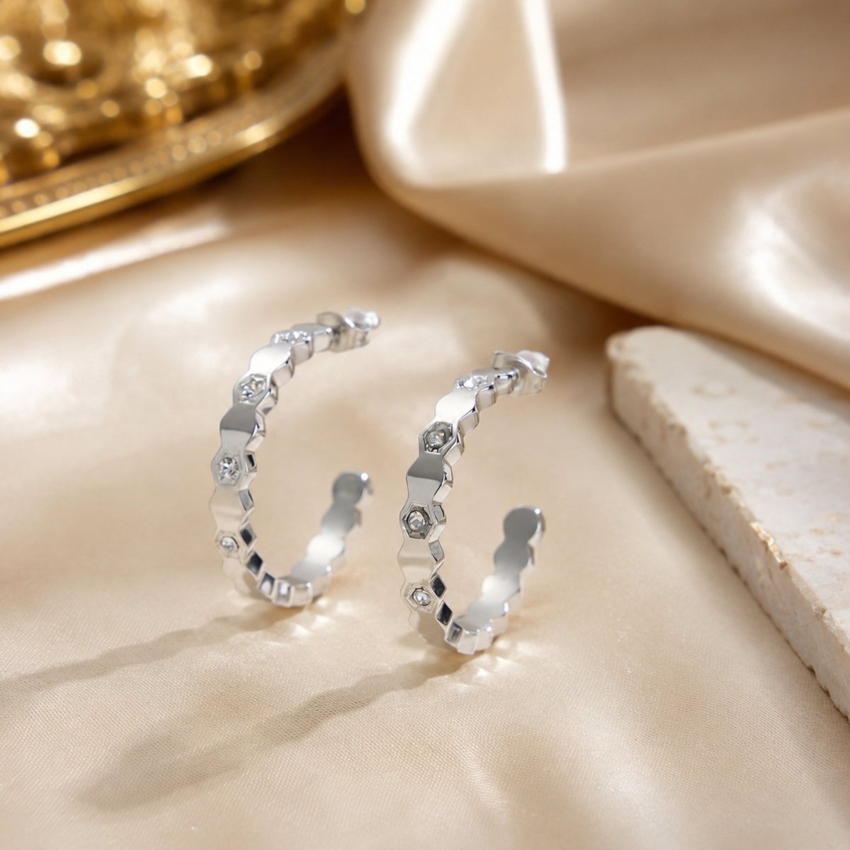 High end silver geometric crystal hoop earrings elegantly styled on glossy champagne silk fabric beside a piece of natural raw stone