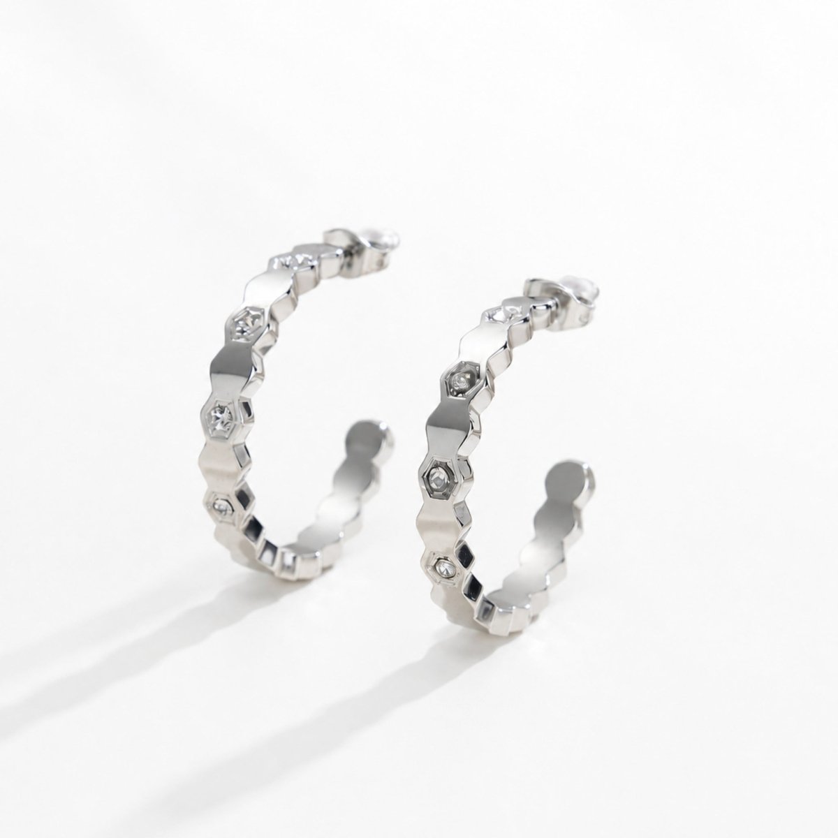 Pair of rhodium silver plated geometric hexagon hoop earrings with clear crystals casting distinct shadows on a bright white background