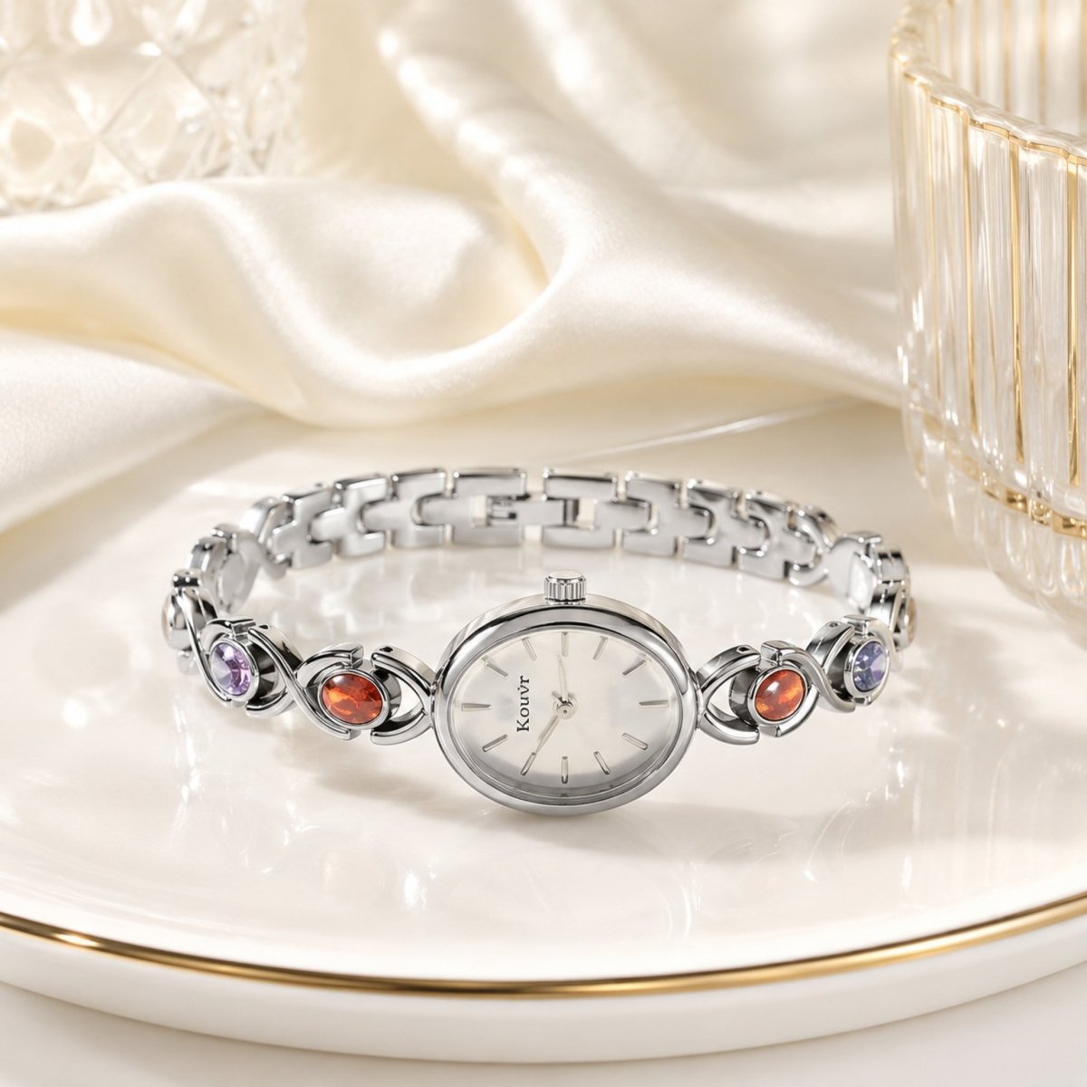 Elegant lifestyle presentation of the silver Kouvr women's jewelry watch lying on a gold-rimmed white display plate alongside luxurious silk draping.