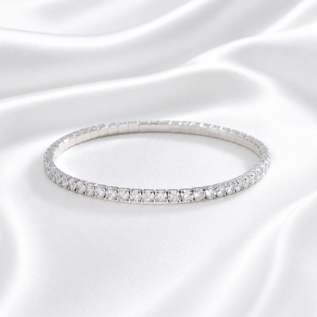 Silver tone clasp-free stretch tennis bracelet lying flat on luxurious white silk fabric showing continuous diamond-like sparkle.
