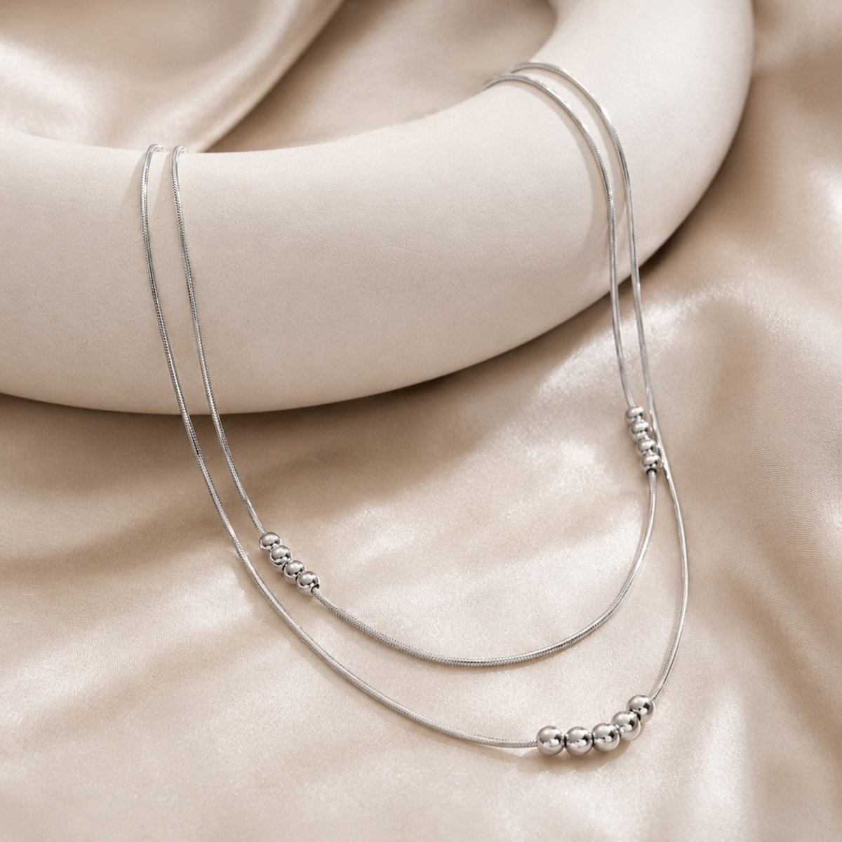 Silver double layered metal bead necklace draped elegantly over a minimalist white ceramic prop against a soft champagne silk background.
