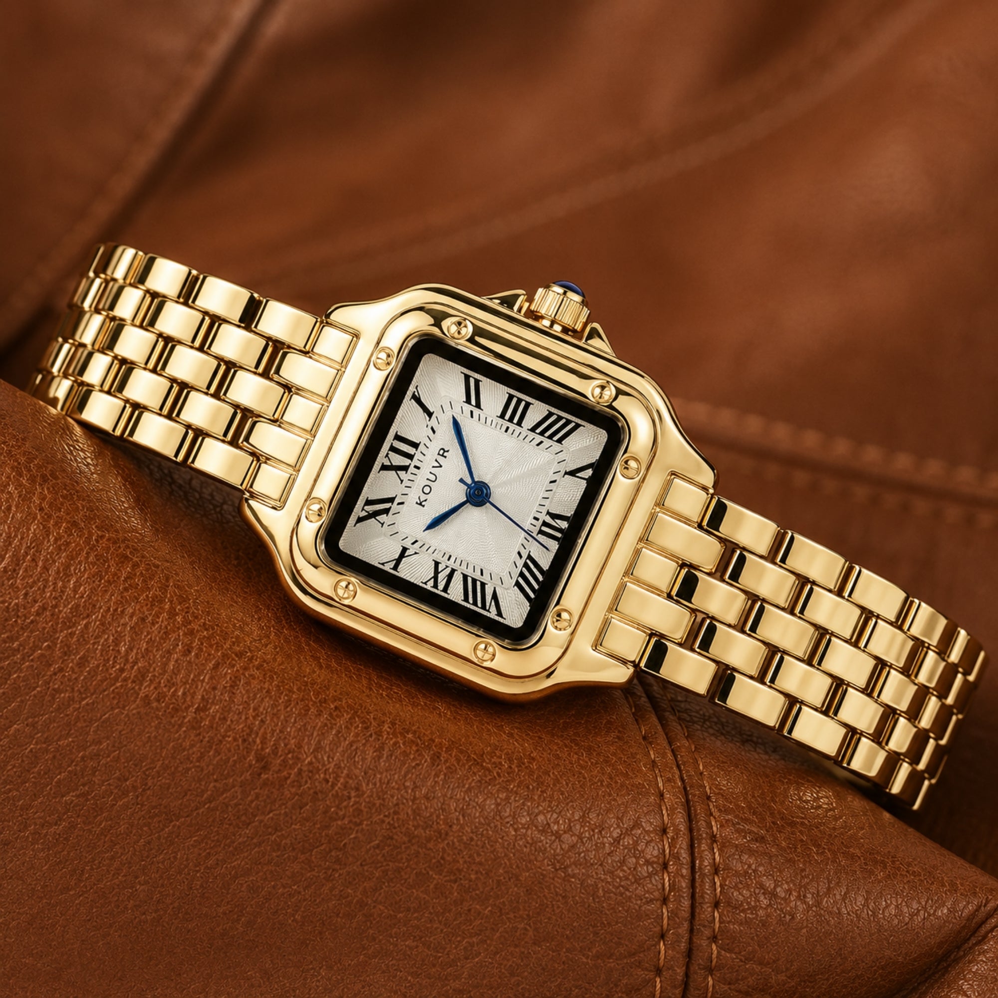 Elegant Kouvr gold-tone women's watch with blue cabochon crown resting flat on a sophisticated brown leather surface.