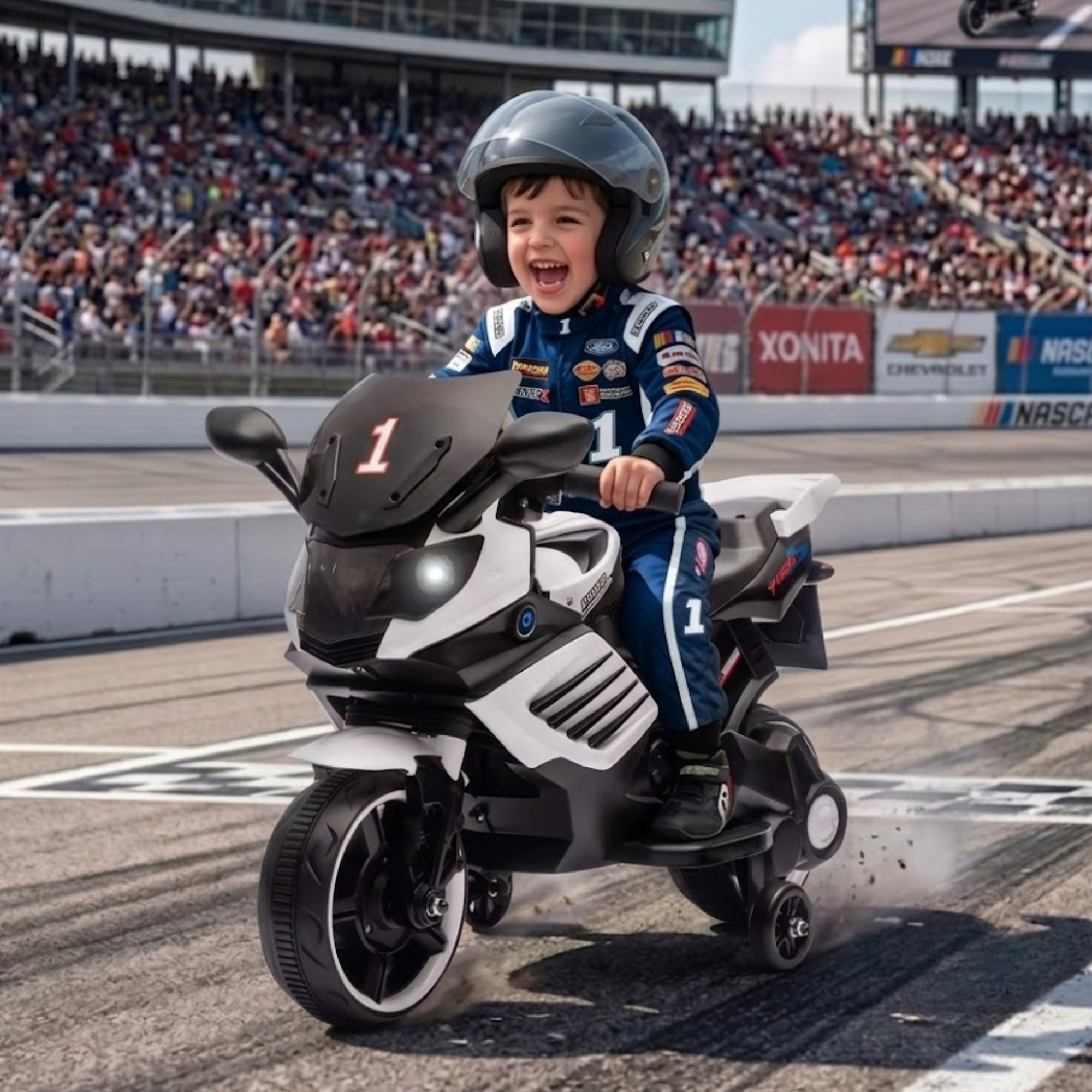 A joyful young boy wearing a gray helmet and blue racing suit, driving a white and black electric ride-on toy motorcycle with training wheels down a racetrack.